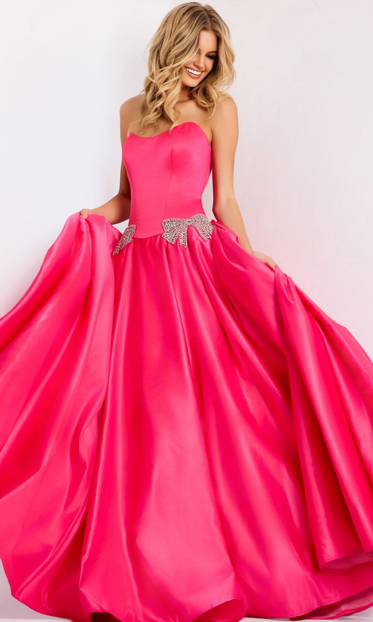 A woman poses against a light background in the fuchsia Jovani 47096 long prom dress, featuring a strapless satin bodice, full skirt, and sparkling rhinestone bow at the waist.
